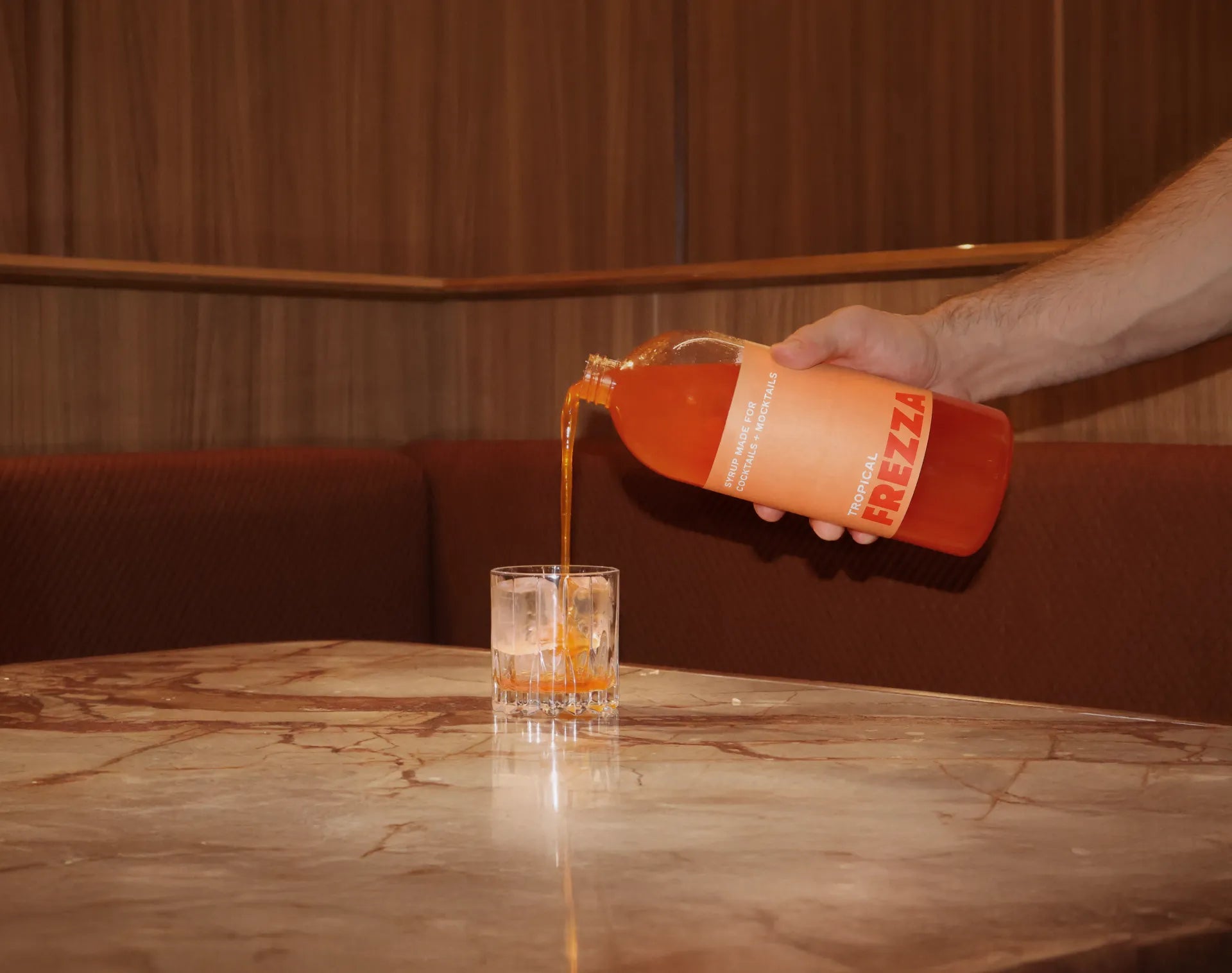 A hand pouring a plastic orange syrup bottle called Frezza Tropical. Orange liquid is poured into a short tumbler glass filled with ice, with the glass set on a marbled tabletop. 